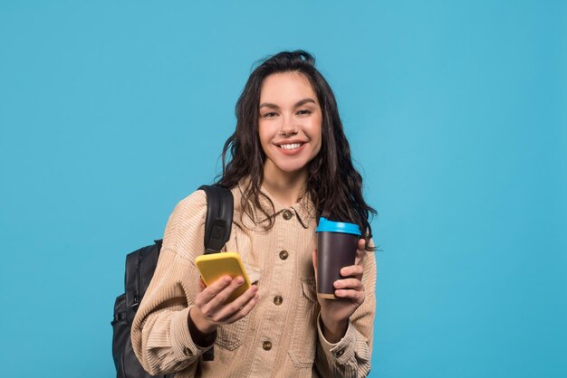 Souriante jeune femme brune européenne avec sac à dos tasse de café et smartphone
