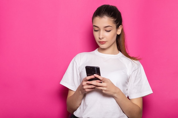 Souriante jeune femme brune fille posant isolée sur le portrait de studio de fond de mur rose pastel. Concept de mode de vie des gens. Maquette de l'espace de copie. Utilisation d'un téléphone portable, saisie d'un message sms