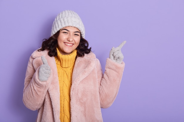 Souriante jeune femme brune pull jaune, manteau de fourrure rose, bonnet gris et mitaines