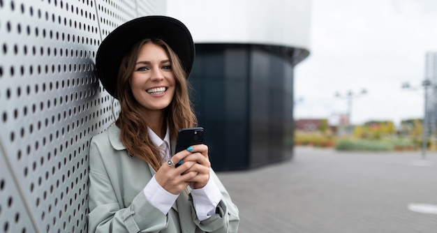 Souriante jeune femme caucasienne d'apparence avec un téléphone portable dans un chapeau à l'extérieur du bureau