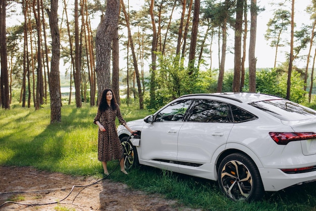 Souriante jeune femme caucasienne branchant le câble électrique dans un véhicule électrique pour recharger sur le parking ensoleillé du centre commercial recadré Concept de mode de vie et d'écologie