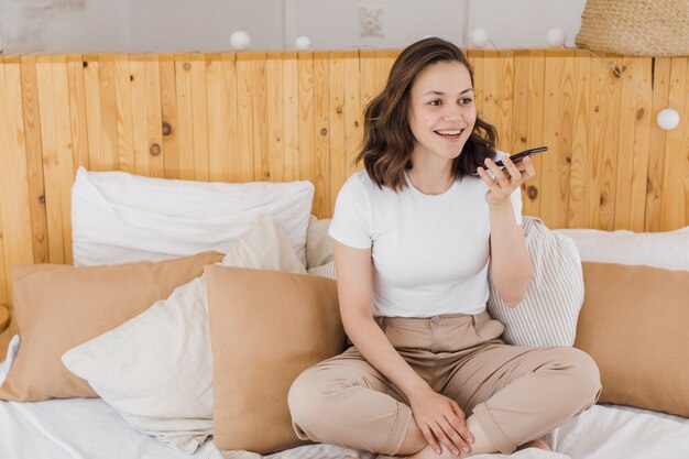 Souriante jeune femme caucasienne enregistrer un message audio numérique sur smartphone dans un appartement moderne