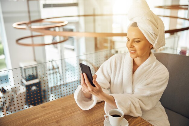 Souriante jeune femme concentrée dans un peignoir tenant son téléphone portable devant elle