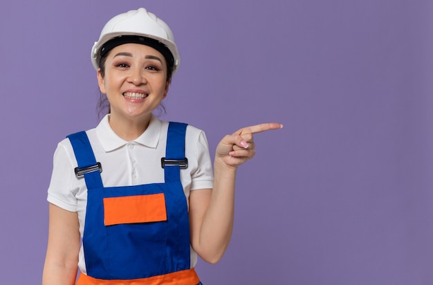 Souriante jeune femme de constructeur asiatique avec un casque de sécurité blanc pointant sur le côté