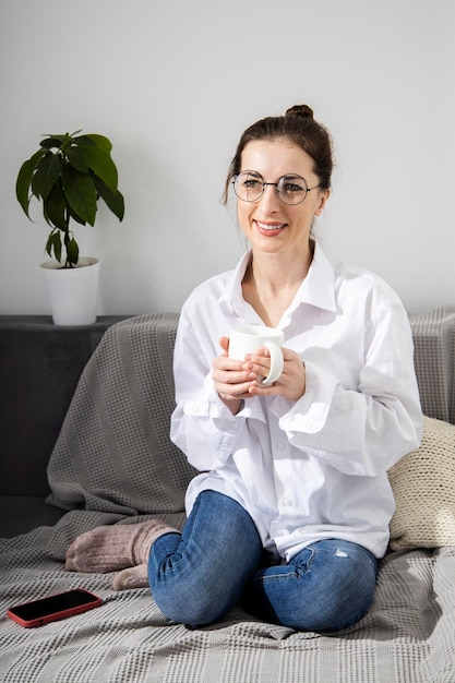 Souriante jeune femme dans des verres avec une tasse de café assis sur un canapé