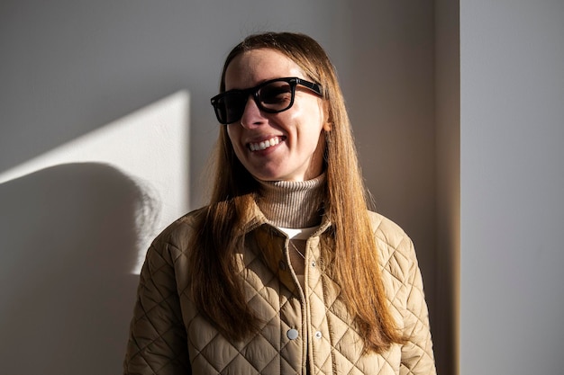 Souriante jeune femme dans une veste à lunettes de soleil contre un mur blanc