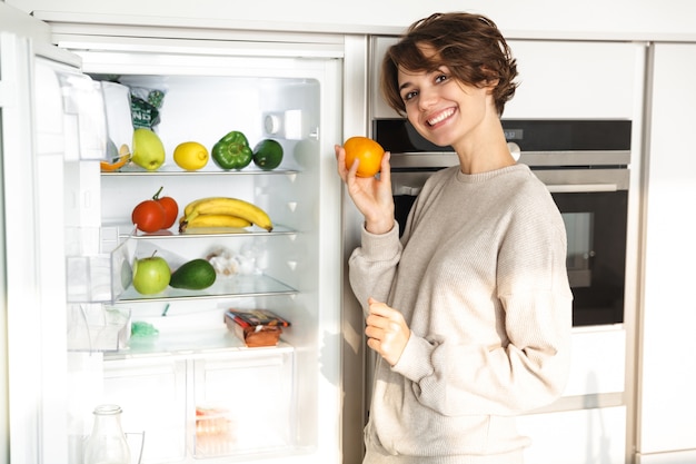 Souriante jeune femme debout au réfrigérateur, tenant orange