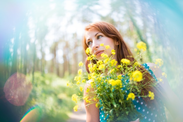 Souriante jeune femme debout, tenant des fleurs jaunes et regardant le ciel dans la forêt le jour d'été ensoleillé. Être sur le concept de la nature