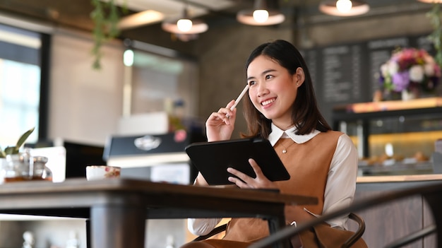 Souriante jeune femme designer tenant une tablette numérique et pensant à son projet