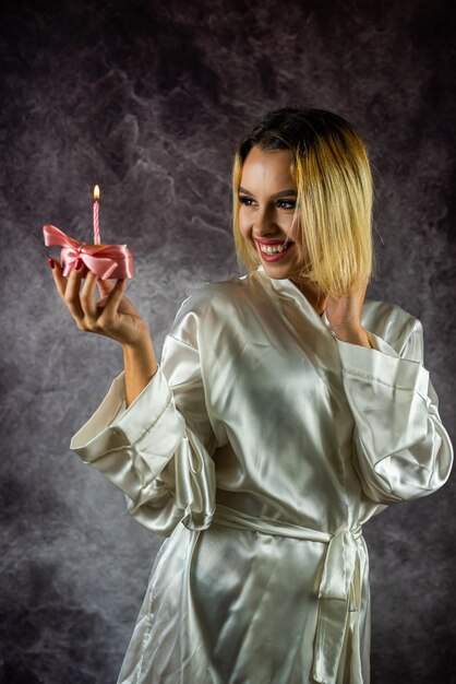 Souriante jeune femme avec du maquillage et en peignoir tenant un petit gâteau d'anniversaire avec une bougie