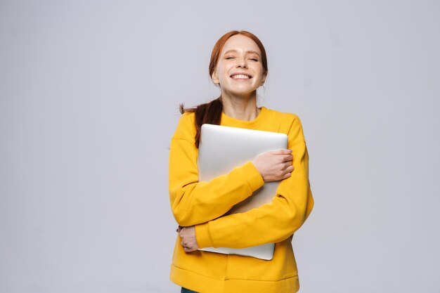 Souriante jeune femme étudiante tenant un ordinateur portable et regardant la caméra sur fond blanc isolé Modèle jolie dame aux cheveux rouges montrant émotionnellement les expressions faciales dans l'espace de copie de studio