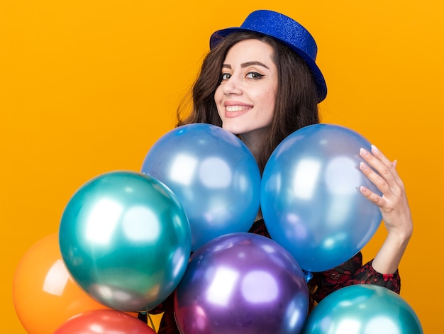 Souriante jeune femme de fête portant un chapeau de fête debout derrière des ballons en touchant un regardant à l'avant isolé sur un mur orange