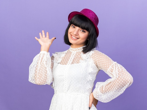 Souriante jeune femme de fête portant un chapeau de fête regardant devant en gardant la main sur la taille montrant une main vide isolée sur un mur violet