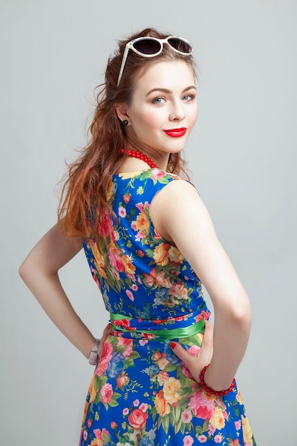 Souriante jeune femme fille rousse dans une robe avec un imprimé floral perles rouges et des lunettes de soleil Fond gris vertical