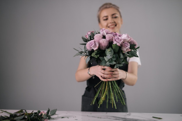 Souriante jeune femme fleuriste portant un tablier tenant un bouquet de fleurs fraîches dans les mains à la table sur fond blanc. Concept de travail avec des fleurs, entreprise florale.