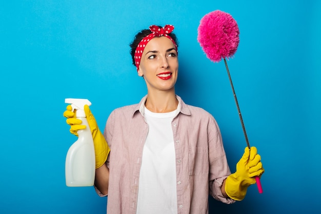 Souriante jeune femme en gants tenant spray et brosse pour nettoyer la poussière sur la surface bleue