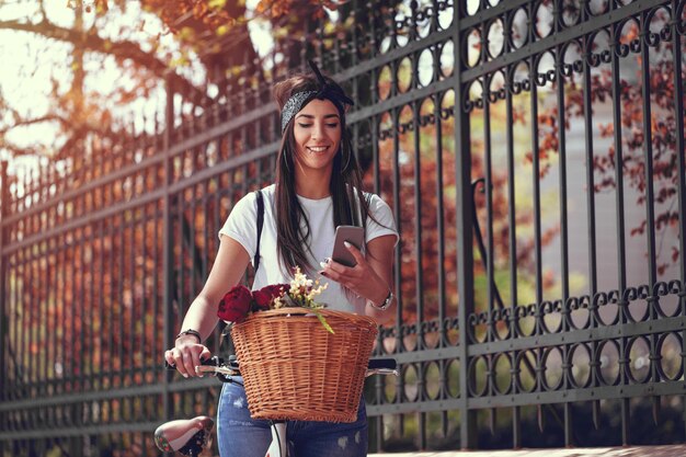 Photo souriante jeune femme lisant un message sur un smartphone dans la rue de la ville, par beau temps, à côté du vélo avec panier de fleurs.