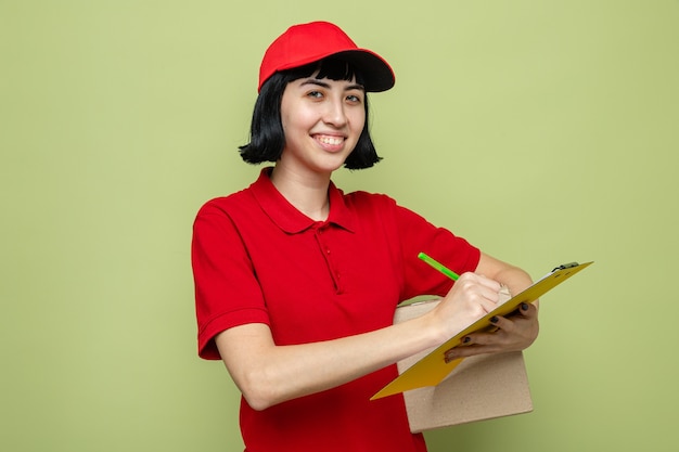 Souriante jeune femme de livraison caucasienne tenant une boîte en carton et écrivant sur le presse-papiers