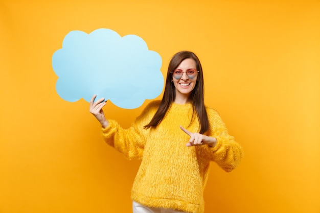 Souriante jeune femme à lunettes coeur pointant l'index sur le bleu vide vide Dire nuage, bulle de dialogue isolée sur fond jaune. Les gens émotions sincères, concept de style de vie. Espace publicitaire.