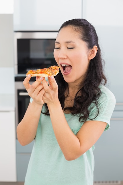 Souriante jeune femme mangeant une tranche de pizza dans la cuisine