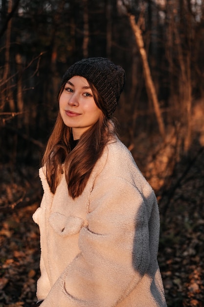 Souriante jeune femme en manteau et chapeau debout dans la forêt d'automne