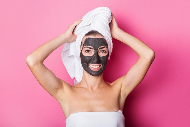 Souriante jeune femme avec masque facial et serviette sur la tête sur fond rose.