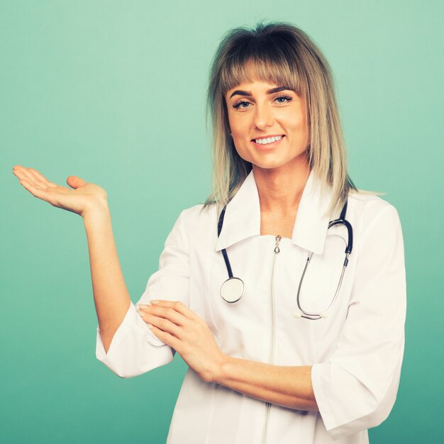 Souriante jeune femme médecin avec un stéthoscope montre quelque chose sur la paume sur un espace bleu