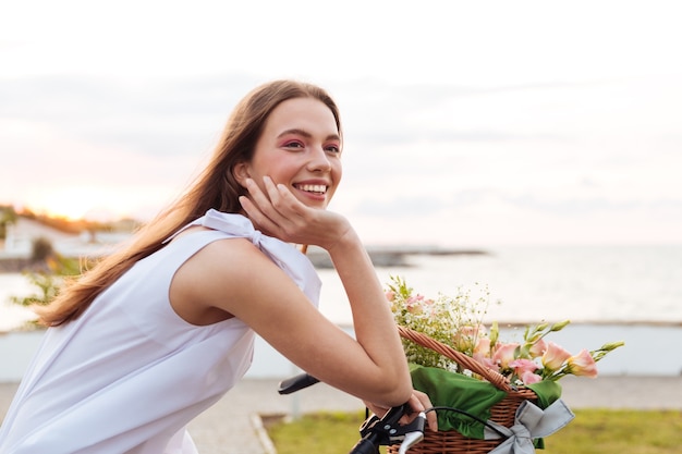 Souriante jeune femme mignonne à vélo avec panier de fleurs en été