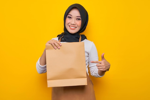 Souriante jeune femme musulmane asiatique barista en tablier de t-shirt blanc travail dans un café Pointant du doigt le sac à emporter en papier vierge maquette isolé sur fond jaune