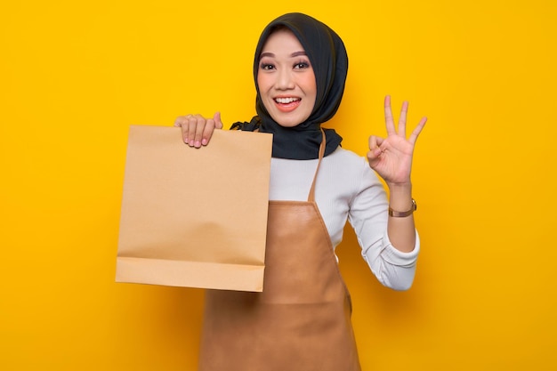 Souriante jeune femme musulmane asiatique barista en tablier de t-shirt blanc travail dans un café tenir un sac à emporter en papier vierge maquette et montrant un geste correct isolé sur fond jaune