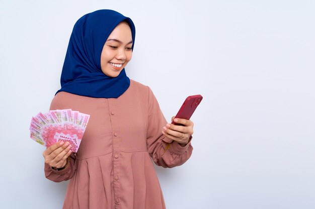 Souriante jeune femme musulmane asiatique en chemise rose à l'aide de téléphone portable et tenant des billets de banque isolés sur fond blanc Concept de style de vie religieux des gens