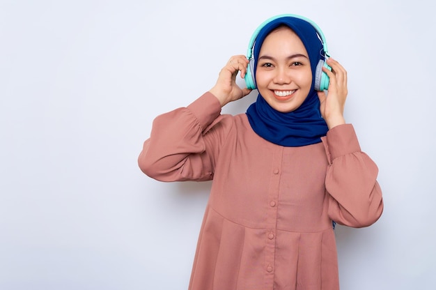 Souriante jeune femme musulmane asiatique en chemise rose écoutant de la musique dans les écouteurs en regardant la caméra isolée sur fond blanc Concept de style de vie religieux des gens