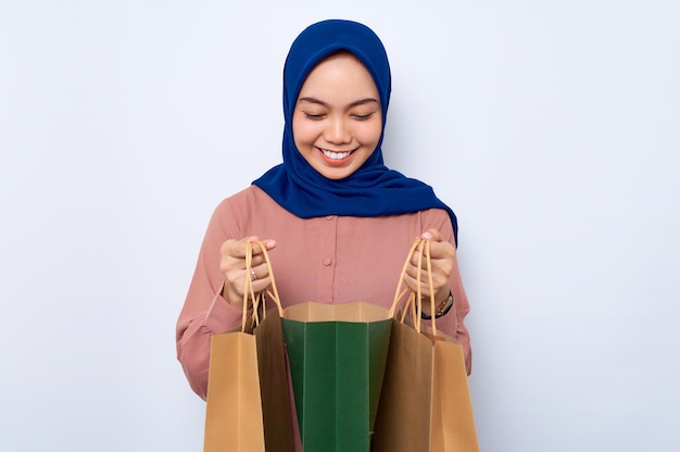 Souriante jeune femme musulmane asiatique en chemise rose ouvrant des sacs d'emballage avec des achats après le shopping isolé sur fond blanc Concept de style de vie des gens