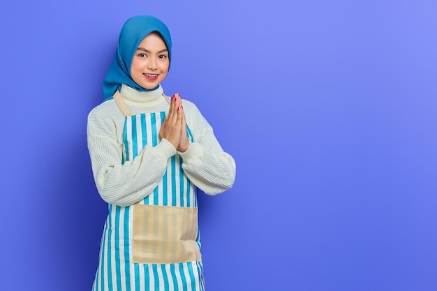 Souriante jeune femme musulmane asiatique dans les années 20 portant le hijab et le tablier signe ouvert pour saluer et saluer les clients isolés sur fond violet Personnes femme au foyer concept de mode de vie musulman