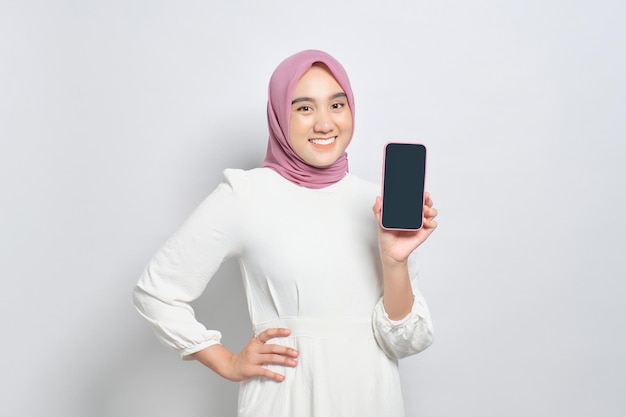 Souriante jeune femme musulmane asiatique montrant un écran vide de téléphone portable recommandant une nouvelle application isolée sur fond blanc