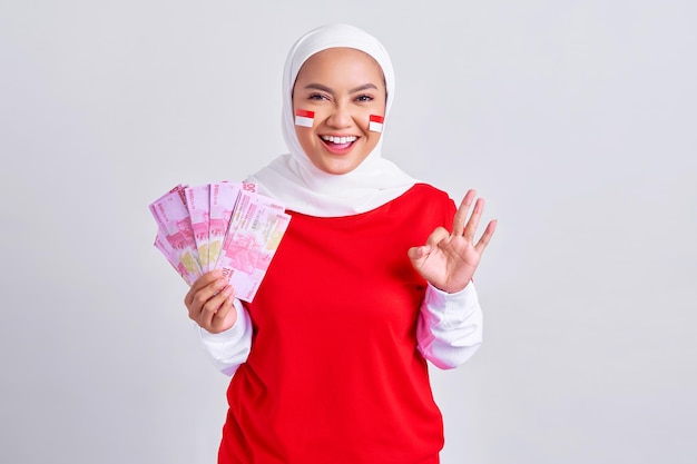 Souriante jeune femme musulmane asiatique en t-shirt blanc rouge tenant de l'argent en espèces en billets de roupie indonésienne montrant un geste correct isolé sur fond blanc Concept de la fête de l'indépendance indonésienne