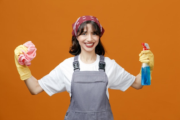 Souriante jeune femme nettoyante portant des gants en caoutchouc uniformes et un bandana montrant un nettoyant et un chiffon en tissu regardant la caméra isolée sur fond orange