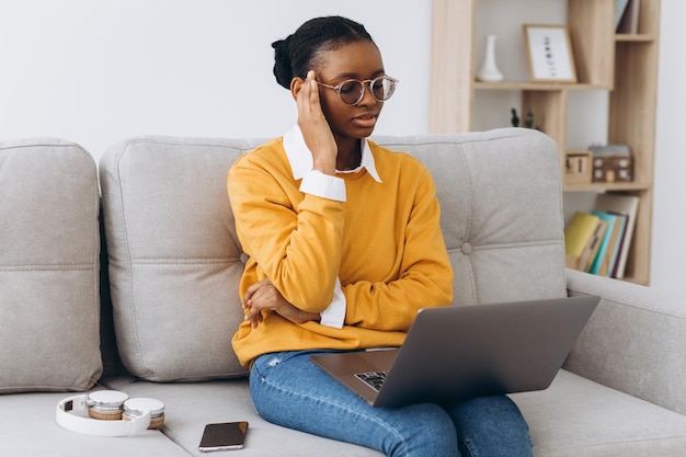 Souriante jeune femme noire en chandail jaune pigiste travaillant à domicile, assise sur un canapé, utilisant un ordinateur portable, copiez l'espace et réfléchissez sérieusement.