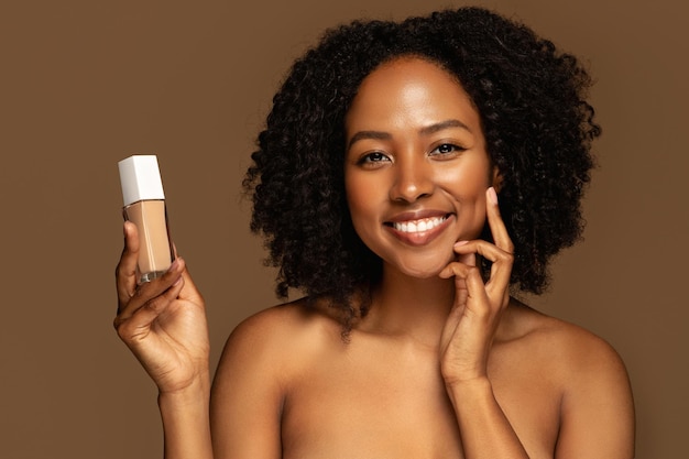 Souriante jeune femme noire séduisante montrant le fond de teint d'un produit de beauté