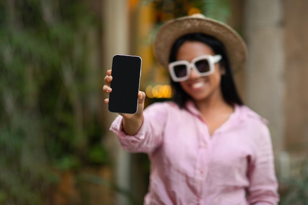 Souriante jeune femme noire touriste en chapeau et lunettes de soleil montre un smartphone avec écran vide profiter de voyager en ville