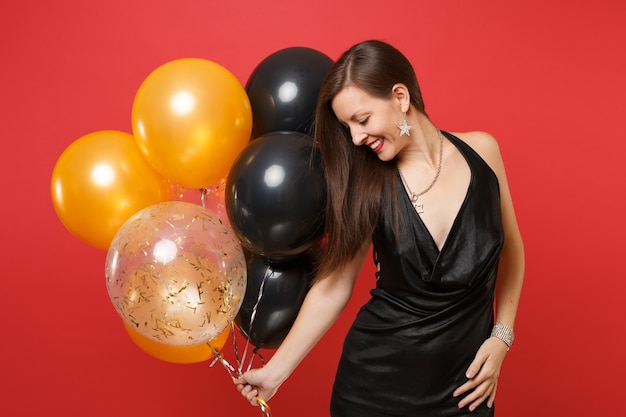 Souriante jeune femme en petite robe noire célébrant, regardant vers le bas, tenant des ballons à air isolés sur fond rouge vif. Saint-Valentin, bonne année, concept de fête de vacances de maquette d'anniversaire.