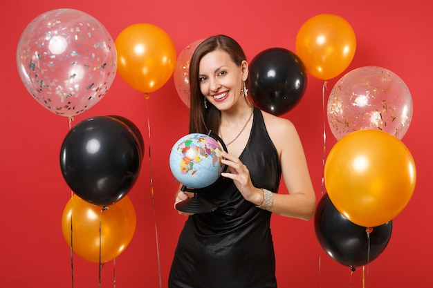 Souriante jeune femme en petite robe noire célébrant et tenant le globe terrestre sur des ballons à air rouge vif. Journée internationale de la femme, bonne année, concept de fête de vacances de maquette d'anniversaire.