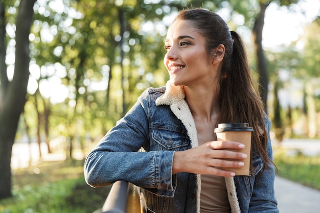 Souriante jeune femme portant une veste assise sur un banc dans le parc, tenant une tasse de café à emporter