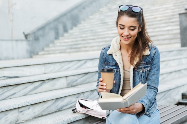 Souriante jeune femme portant une veste assise sur un banc à l'extérieur, livre de lecture, tenant une tasse de café à emporter