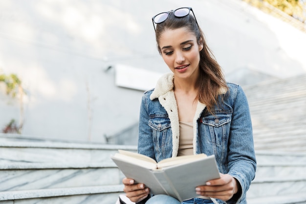 Souriante jeune femme portant une veste assise sur un banc à l'extérieur, livre de lecture