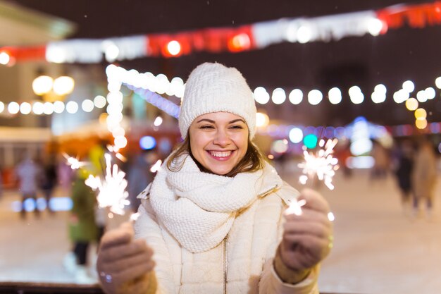 Souriante jeune femme portant des vêtements tricotés d'hiver tenant un cierge magique à l'extérieur sur fond de neige