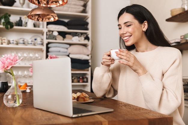 Souriante jeune femme prenant son petit déjeuner assis à la cuisine et à l'aide d'un ordinateur portable