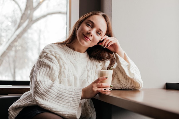 Souriante jeune femme en pull assis au café et boire du café en regardant la caméra à l'intérieur