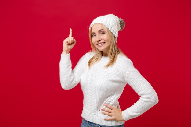 Photo souriante jeune femme en pull blanc, chapeau isolé sur fond rouge portrait en studio. mode de vie sain, émotions des gens, concept de saison froide. maquette de l'espace de copie. pointant l'index vers le haut.