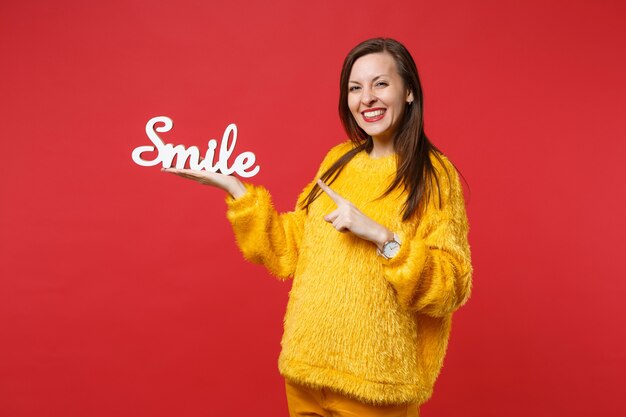 Souriante jeune femme en pull de fourrure jaune pointant l'index de côté sur les lettres de mots en bois sourire isolé sur fond rouge en studio. Les gens émotions sincères, concept de style de vie. Maquette de l'espace de copie.